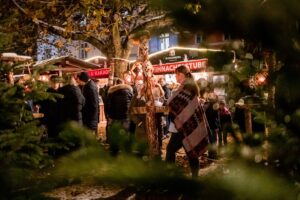 Stimmungsvoller Hüttenzauber mit Menschen, die in einem Winterwald um Holz-Stehtische stehen, sich amüsieren und Glühwein trinken.