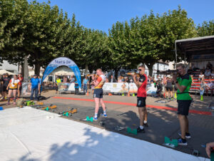 Drei Teilnehmer des Finallaufs beim 6. Citybiathlon Holzminden 2024 absolvieren das Stehendschießen. Im Hintergrund schaut das Publikum stehend und sitzend auf der Zuschauertribühne zu.