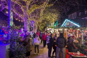 Impressions-Foto von Menschen, die über den Weihnachtsmarkt schlendern. Im Hintergrund sieht man Hütten. Insgesmt ist alles stimmungsvoll beleuchtet.