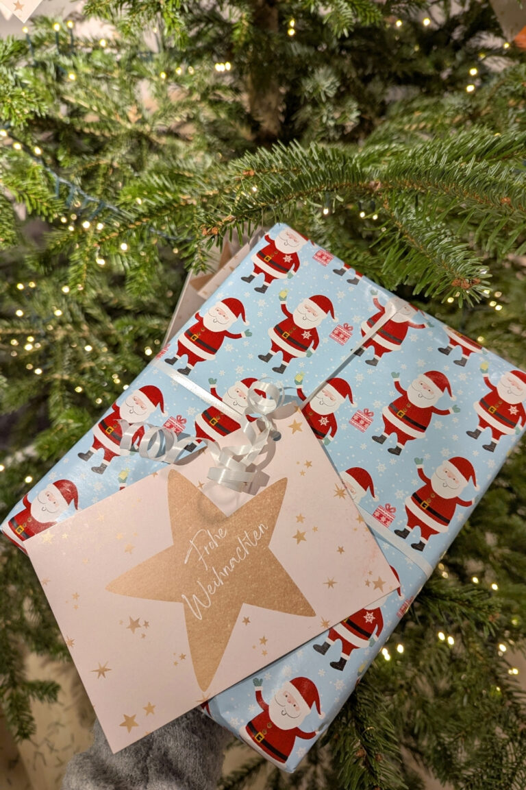 Wunschsternkarte an einem in Weihnachtspapier verpacktem Geschenk befestigt mit einem beleuchteten Tannenbaum im Hintergrund