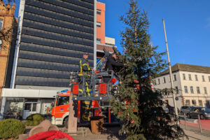 Bürgermeister Christian Belke steht auf einer Drehleiter der Feuerwehr und schmückt den "Little Ugly Christmas Tree" mit roten Christbaumkugeln.