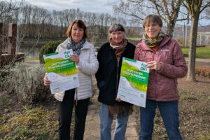 Von links: Marion Lampe, Elisabeth Kilian und Ruth Koßmann stehen im Duftgarten mit der Weser im Hintergrund und halten das Plakat zum duftenden Stadtrundgang 2026 in die Kamera.