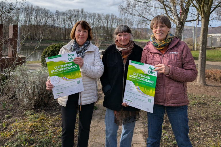 Von links: Marion Lampe, Elisabeth Kilian und Ruth Koßmann stehen im Duftgarten mit der Weser im Hintergrund und halten das Plakat zum duftenden Stadtrundgang 2026 in die Kamera.