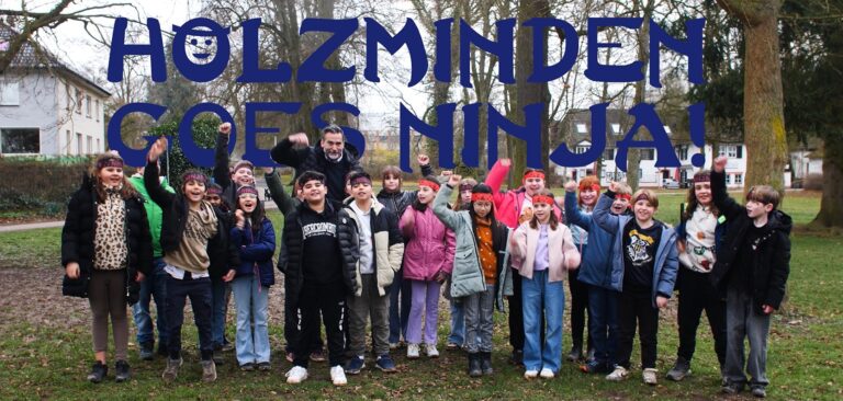 Eine Schulklasse mit Ninja-Stirnbändern und Bürgermeister Christian Belke stehen im Park an den Teichen, wo der Ninja-Parcours entstehen soll.