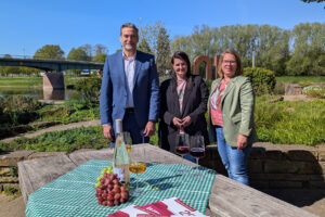 Bürgermeister Belke, Stadtmarketing-Geschäftsführerin Michaela Reinecke-Koller und Inga Schaper posieren vor einem mit Wein, gefüllten Weingläsern und Weintrauben dekorierten Tisch bei strahlendem blauen Himmel und Sonnenschein im Duftgarten.