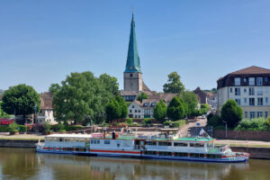 Das Fahrgastschiff "Höxter" der Flotte Weser liegt am Weserkai in Holzminden mit dem Duftgarten und Lutherkirche im Hintergrund.