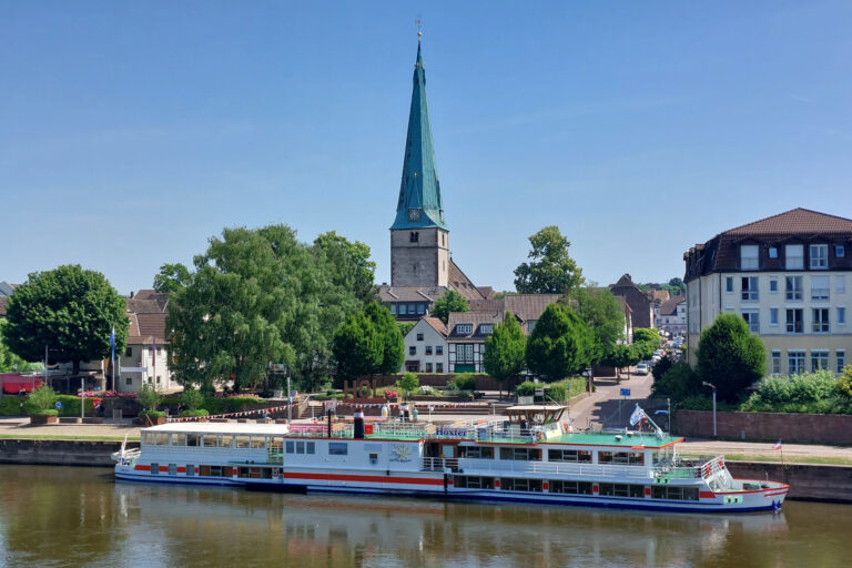 Das Fahrgastschiff "Höxter" der Flotte Weser liegt am Weserkai in Holzminden mit dem Duftgarten und Lutherkirche im Hintergrund.