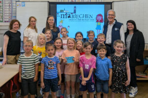 Die Kinder der 1. Klasse der Katholische Grundschule Holzminden gemeinsam mit Isabell Adam vom Stadtmarketing, Schulliterin Stefanie Roland, Autorin Nina Schiefelbein, Erich Werner, Präsident des Lions Club Holzminden und Michaela Reinecke-Koller, Geschäftsführerin Stadtmarketing Holzminden GmbH im Klassenraum vor der Tafel, die das Mariechentitelbild zeigt.