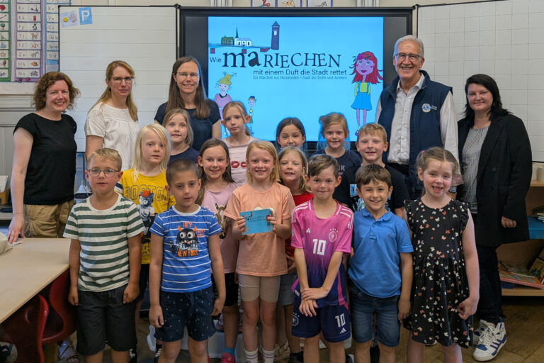 Die Kinder der 1. Klasse der Katholische Grundschule Holzminden gemeinsam mit Isabell Adam vom Stadtmarketing, Schulliterin Stefanie Roland, Autorin Nina Schiefelbein, Erich Werner, Präsident des Lions Club Holzminden und Michaela Reinecke-Koller, Geschäftsführerin Stadtmarketing Holzminden GmbH im Klassenraum vor der Tafel, die das Mariechentitelbild zeigt.