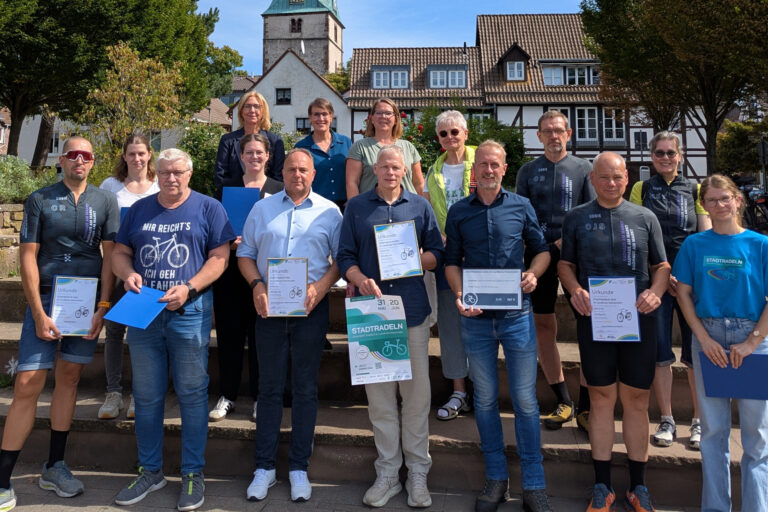 Die Organisatoren, Sponsoren und ausgezeichneten Radfahrerinnen und Radfahrer des STADTRADELN 2026 im Landkreis Holzminden posieren am Weserkai vor der Treppe des Duftgartens mit diesem und der Lutherkirche im Hintergrund.