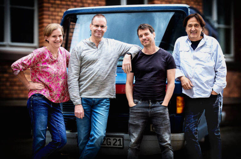 Die Band posiert draußen vor einem alten Wagen.
