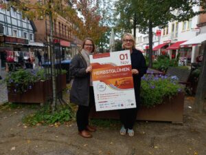 Inga Schaper vom Stadtmarketing und Bianca Kleeschulte vom Werbekreis stehen am Eingang zur Fußgängerzone in der Oberen Straße und halten ein Veranstaltungsplakat in Kamera.